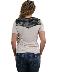 A woman viewed from behind wearing a short-sleeve t-shirt with a beige body and a dark green and black camouflage yoke across the shoulders, paired with dark blue jeans.