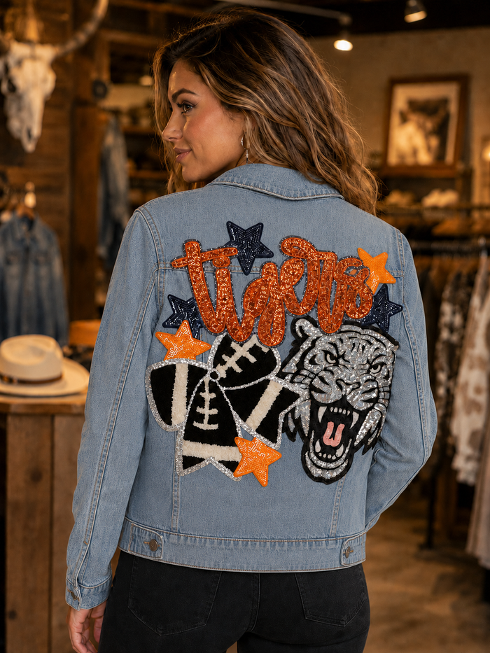 A woman wearing a light blue denim jacket with a large, colorful back patch featuring the word 'Twizt' in orange glitter, football, stars, and a tiger head, photographed in a rustic clothing store.