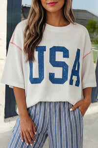 Person wearing a white sweater with 'USA' printed in blue and striped pants outdoors.
