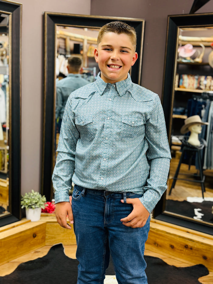 A young boy smiling in a retail store, wearing a light blue and white patterned western-style button-down shirt with blue jeans.