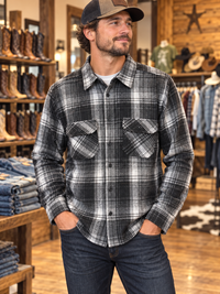 A sexy man wearing a black and gray plaid flannel button-down shirt with two chest pockets, paired with dark jeans and a brown baseball cap, standing inside a rustic western-style clothing store.