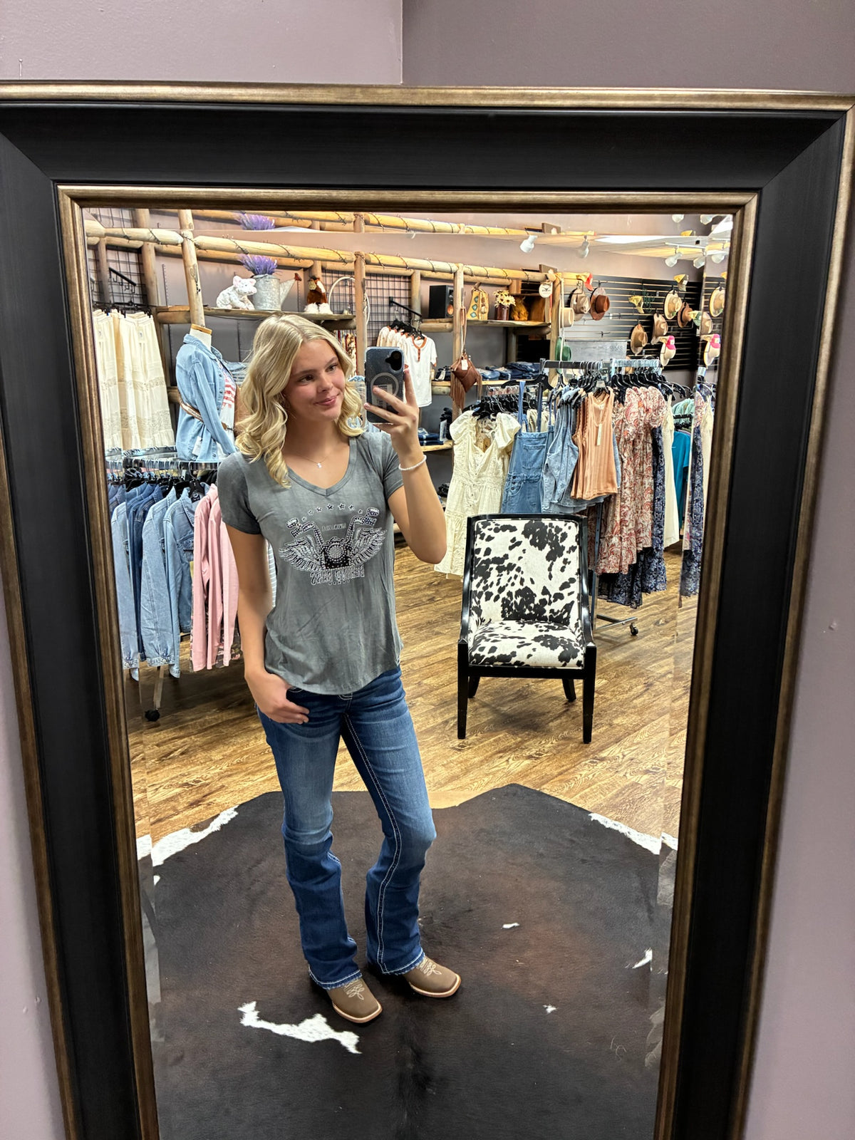 A woman takes a mirror selfie in a clothing store, wearing a gray t-shirt with a graphic print, blue jeans, and tan shoes. The photo is framed by a large black mirror with a gold-toned inner border.