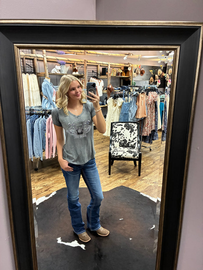 A woman takes a mirror selfie in a clothing store, wearing a gray t-shirt with a graphic print, blue jeans, and tan shoes. The photo is framed by a large black mirror with a gold-toned inner border.