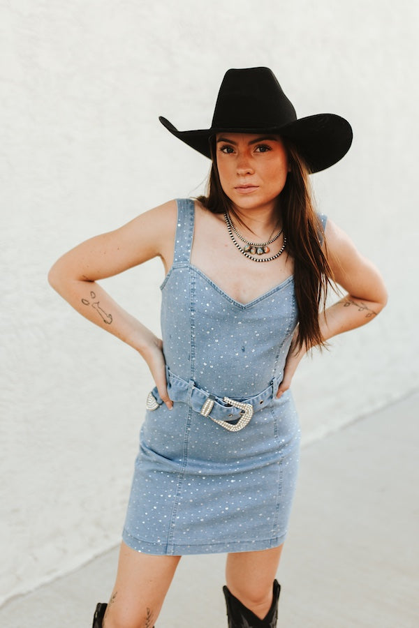 woman in cowgirl hat wearing rhinestone dress with belt and boots up against a white wall