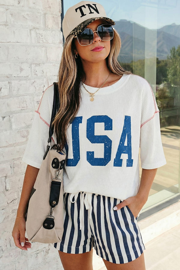Woman wearing a white t-shirt with 'USA' print, striped shorts, and a cap outdoors.