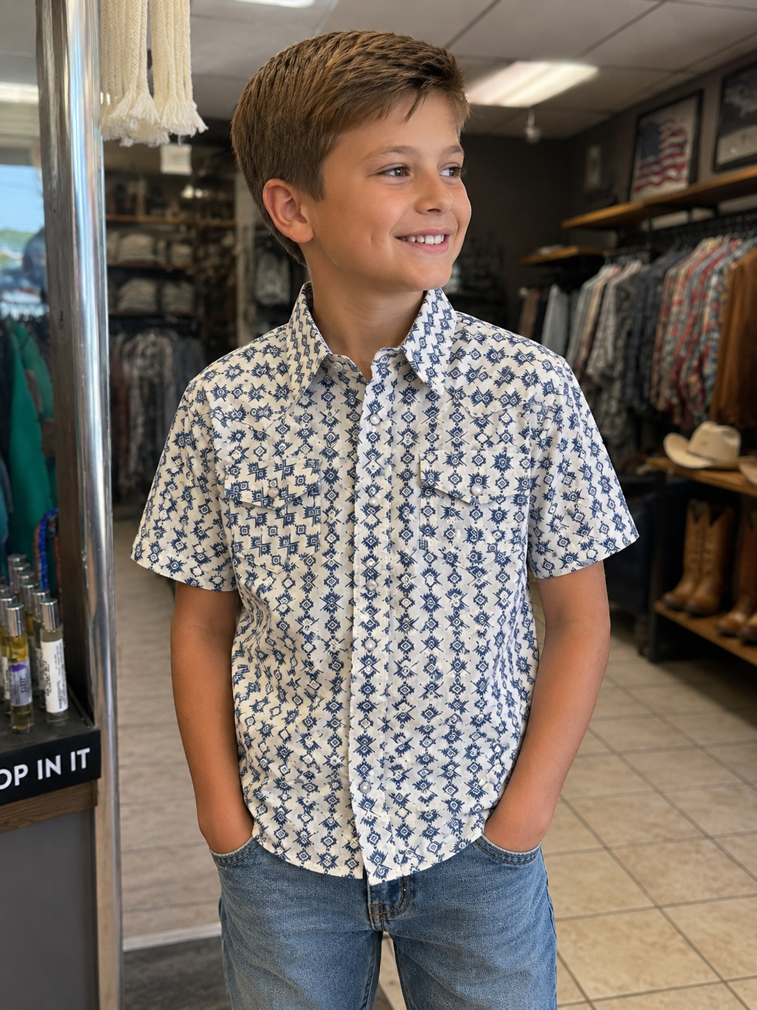 A young boy smiling in a clothing store, wearing a short-sleeve button-down shirt with a blue and white geometric pattern and blue jeans.