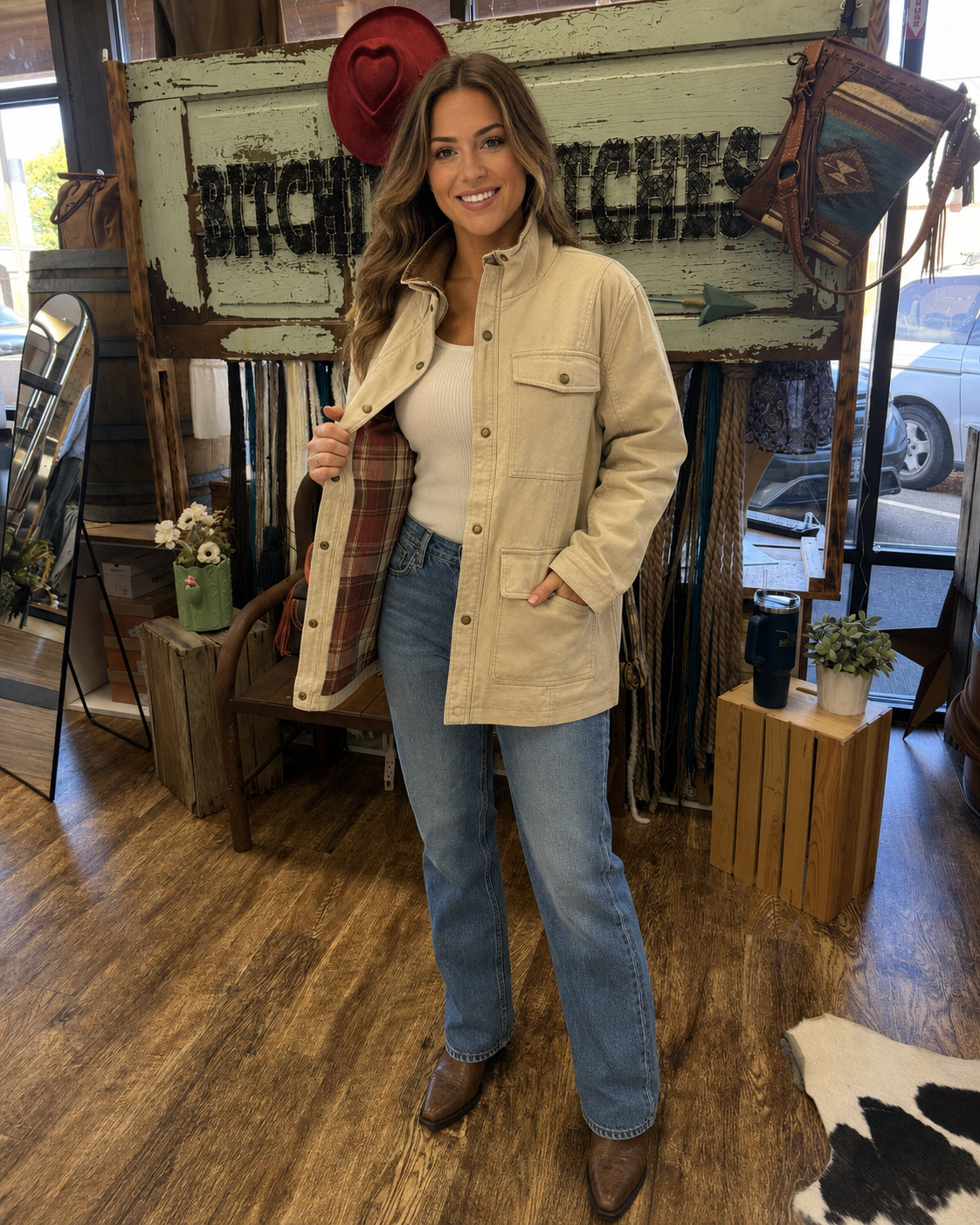 A woman models a beige button-up utility jacket with a red and green plaid lining, styled with blue jeans and brown boots in a rustic shop setting.